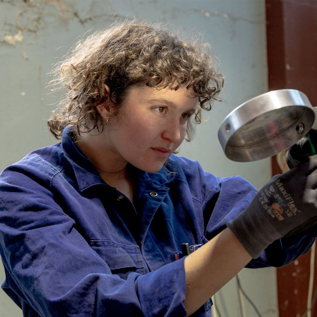Photo of women in blue overalls with curly hair holding a silver fixture, wearing gloves. 